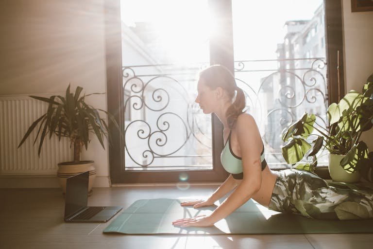 A woman performing cobra pose yoga indoors next to a window with sunlight streaming in.