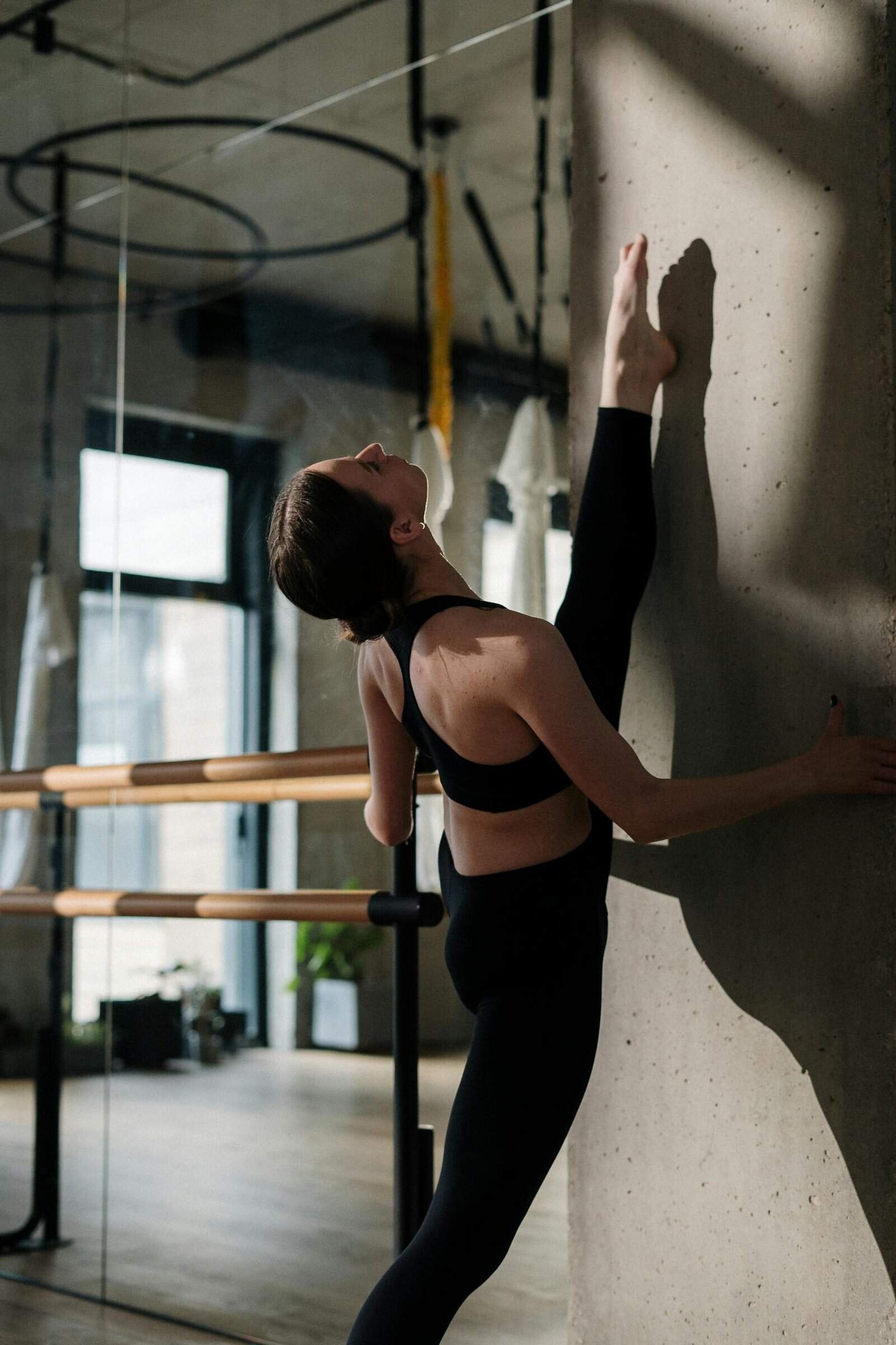 A woman stretches against a concrete wall in a bright yoga studio, demonstrating flexibility.