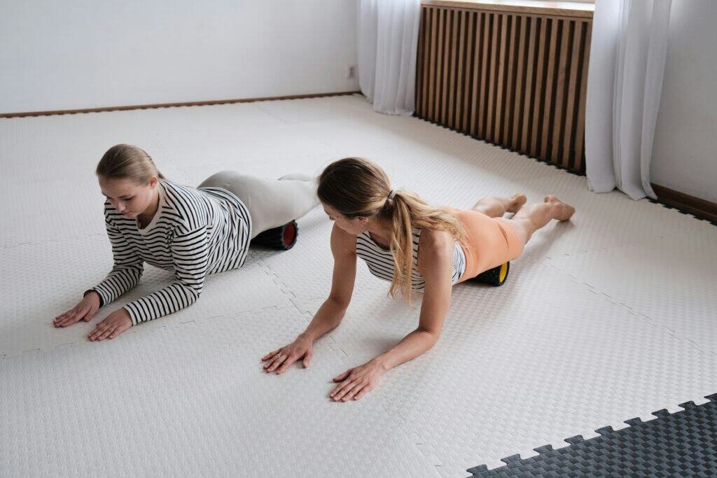 Two women doing back stretches on a white mat in a home gym.