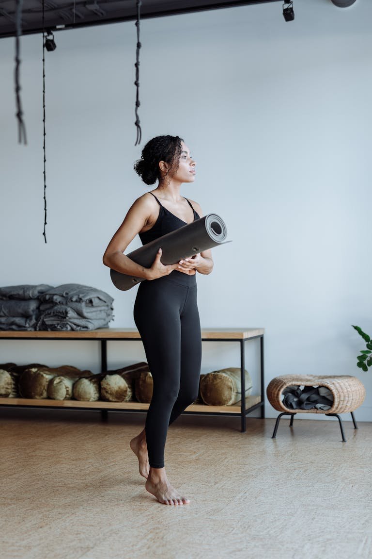 Black woman standing in a yoga studio holding a yoga mat, showcasing activewear and fitness lifestyle.