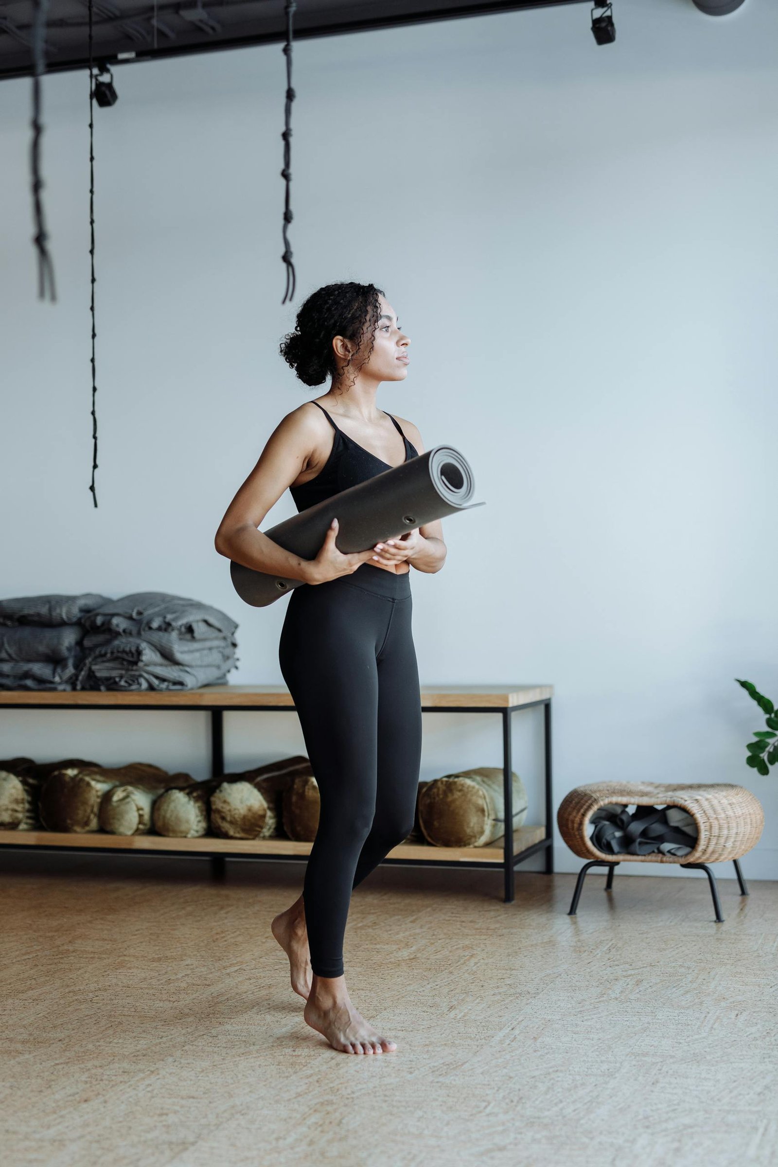 Black woman standing in a yoga studio holding a yoga mat, showcasing activewear and fitness lifestyle.