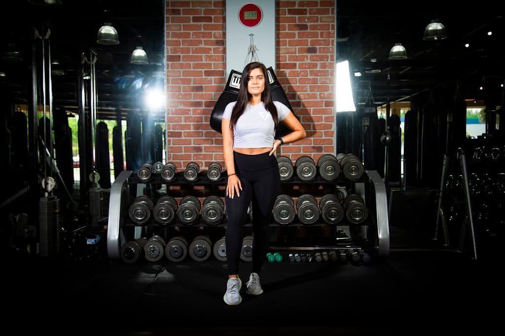 Confident young woman standing in a gym, surrounded by dumbbells, exuding fitness motivation.