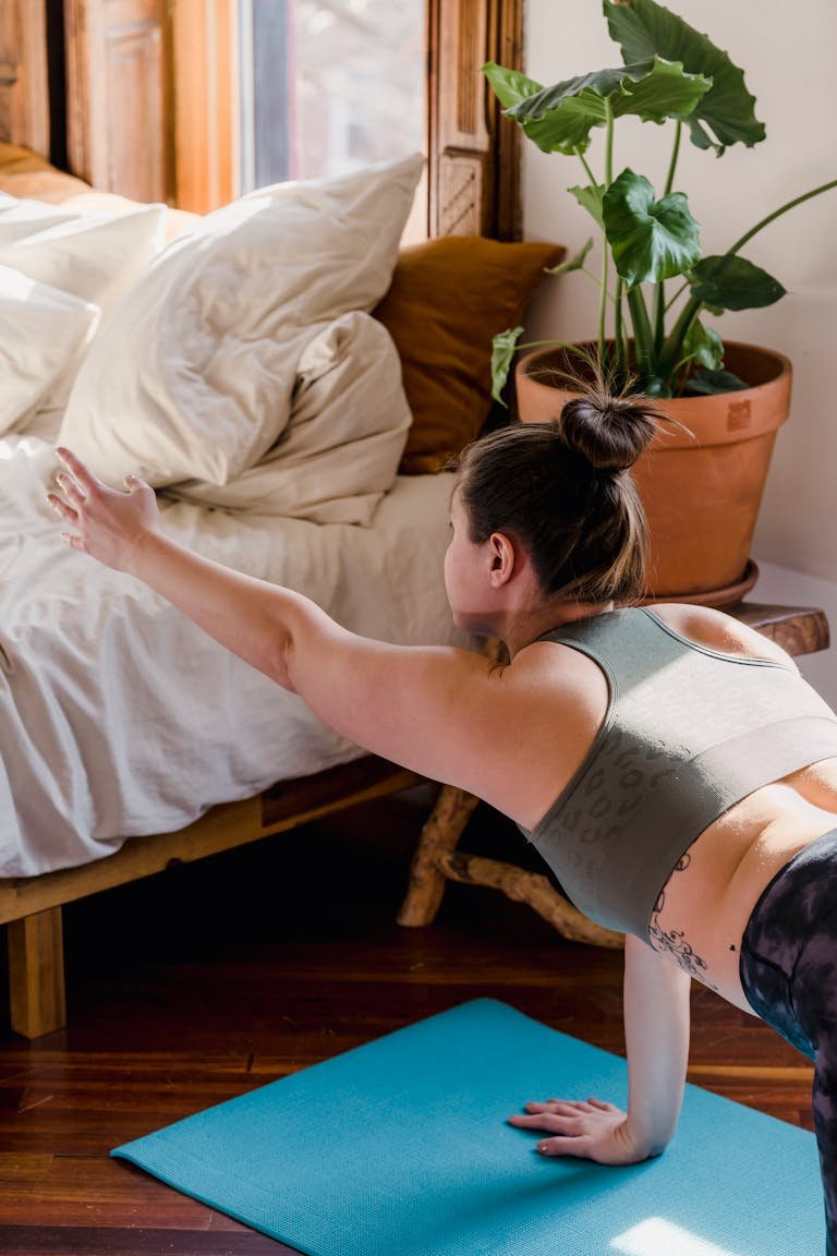 Woman doing yoga stretch on blue mat near bed with indoor plant, embracing morning light.